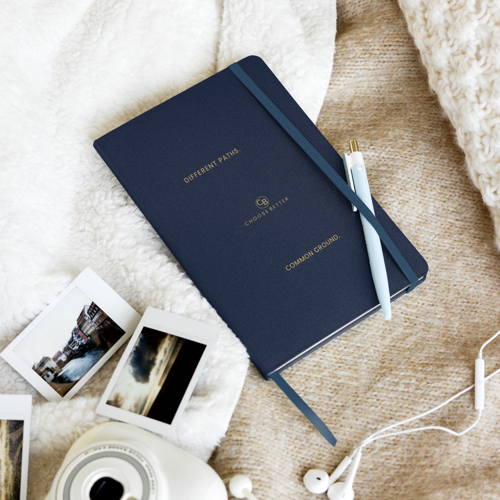 Dark blue notebook with Different Paths. Common Ground. Choose Better branding on a textured surface with photos and earphones.