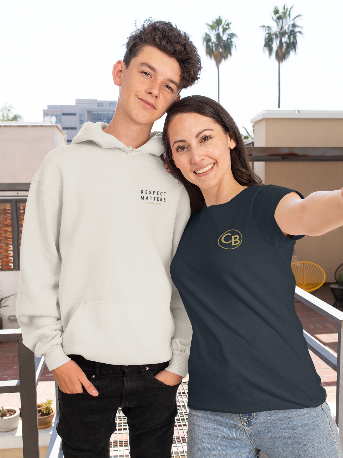 Two people wearing hoodies with logos, standing on a balcony with palm trees in the background.